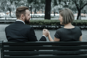 A photo of a man with a beard and a woman with short hair sitting on a bench. The man is wearing a suit and the woman is wearing a dress. They are holding hands. The background is a park with trees.